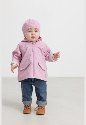 Toddler standing indoors wearing a pink knit hat, pink jacket, rolled-up jeans, and brown shoes on a light floor with white background.
