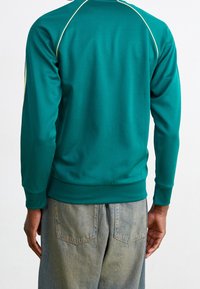 Grön sweatshirt med långa ärmar och ribbade muddar, med gul piping längs axelsömmarna, ihop med blå denimbyxor.