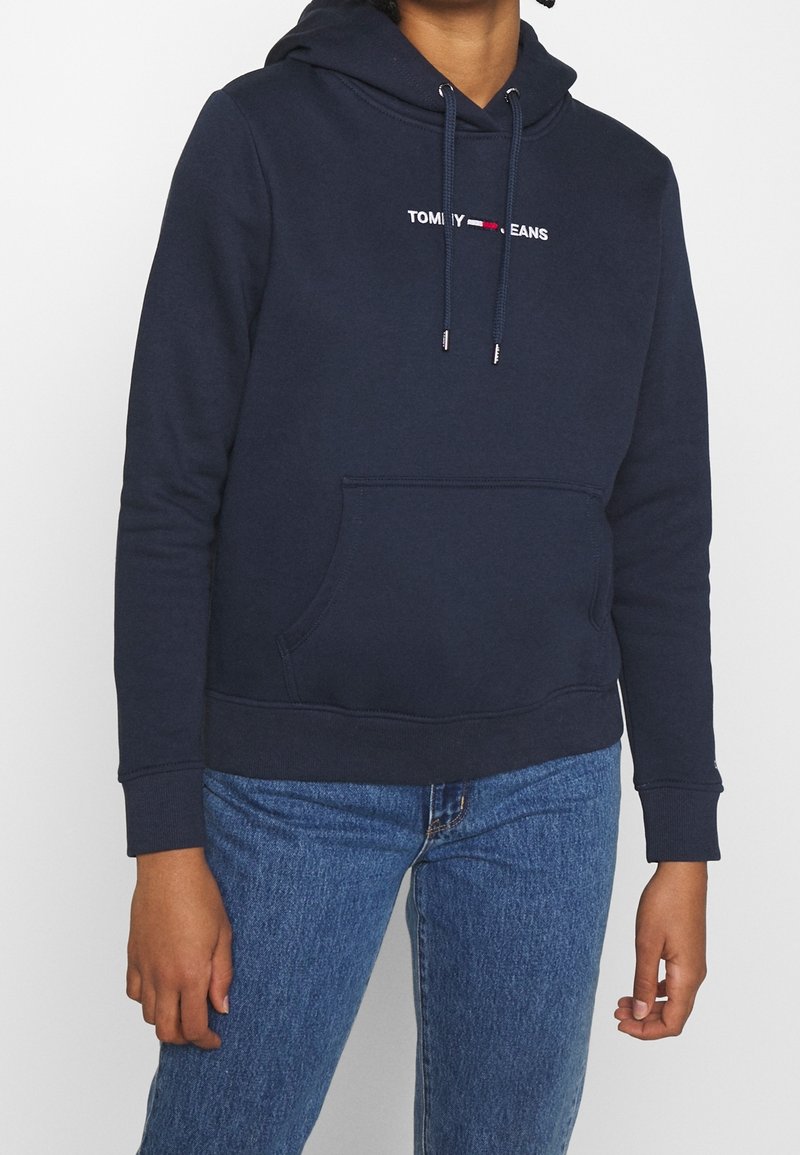 Hoodie bleu marine en mélange de coton doux. Présente une capuche avec cordon de serrage, une poche frontale et un logo "TOMMY JEANS" brodé sur la poitrine.