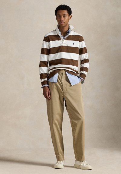 Uomo in piedi che indossa un pullover a righe marroni e bianche, camicia blu con colletto, pantaloni beige e scarpe da ginnastica bianche su sfondo uniforme.