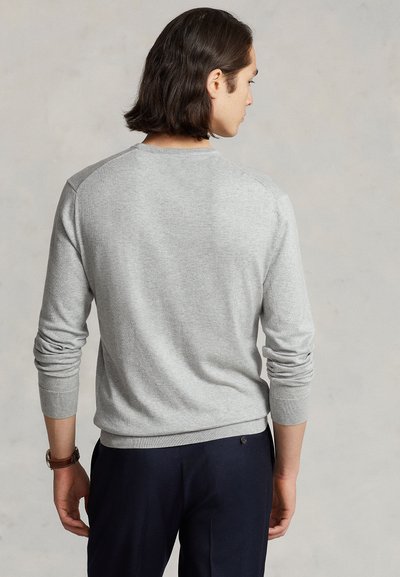 Uomo con capelli scuri fino alle spalle che indossa un maglione grigio chiaro a maniche lunghe e pantaloni scuri, visto da dietro su sfondo uniforme.