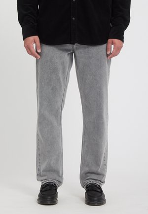 Homme portant un jean droit gris clair, des chaussures noires et une chemise noire à manches longues, debout devant un fond uni.