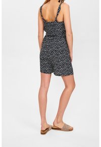 Žena oblečená v černém květinovém bezramenním overal s nastavitelnými ramínky a béžovými sandály na volné špičce, stojící na bílém pozadí.