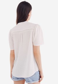 Witte blouse met korte mouwen, voorzien van een gestructureerd patroon, kantdetails op de rug en een relaxte pasvorm, gecombineerd met lichte denimshorts.