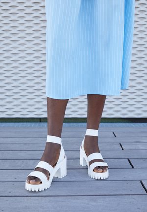 Persoon die witte, stevige hoge hakken sandalen met enkelbandjes draagt en een lichtblauwe rok met fijne strepen, staand op een houten dekvloer.