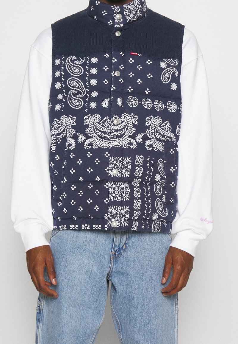 Veste sans manches bleu marine à imprimé patchwork bandana avec boutons-pression portée sur une chemise blanche à manches longues et un jean bleu clair.