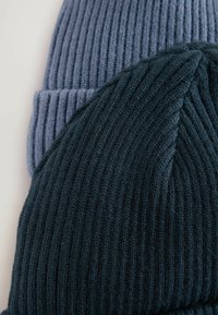 Deux bonnets en tricot ribbée, l'un en bleu doux et l'autre en vert foncé, mettant en valeur un tissu épais et texturé avec un design à revers.