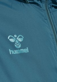 Gros plan d’un tissu turquoise avec le logo « hummel » et un dessin d’abeille stylisé près d’une couture de fermeture éclair assortie.