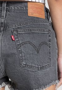 Shorts in denim grigio con orlo sfilacciato, dotati di una tasca posteriore con design cucito e un'etichetta in pelle marrone in vita. Etichetta del marchio rossa visibile.
