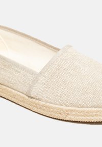 Beige espadrille with a textured fabric upper, elasticized sides, and a woven jute sole, featuring a low-profile design.