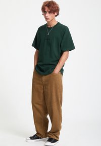 Zielona, przerośnięta koszulka z subtelnym logo, połączona z brązowymi spodniami z korduroju. Czarne sneakersy dopełniają swobodny look. Gładka faktura materiału.