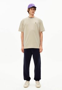 Beige katoenen T-shirt met een klein groen logo, gecombineerd met loszittende marineblauwe broek en een paarse pet. Schoenen zijn beige met een gestructureerd ontwerp.