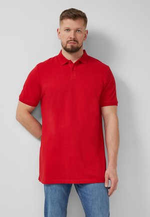 Man met korte baard, gekleed in een effen rood poloshirt en blauwe spijkerbroek, staand tegen een grijze muur.