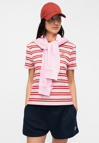 Κοντομάνικο πουκάμισο με κόκκινες και λευκές ρίγες, ροζ πουλόβερ δεμένο γύρω από το λαιμό, ναυτικό σορτς και κόκκινο καπέλο. Βαμβακερό μείγμα, casual σχεδιασμός.