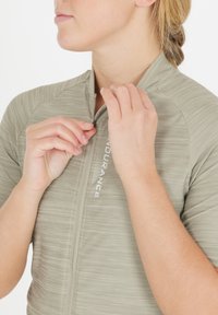 Maillot de cyclisme gris clair avec une finition texturée, doté d'une fermeture éclair à l'avant et du mot "ENDURANCE" imprimé sur la poitrine. Gros plan de mains en train de zipper.