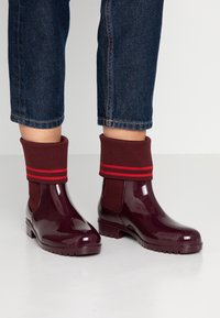 Botas de lluvia de corte bajo en rojo oscuro brillante con puños de punto doblados que presentan dos rayas rojas, usadas con unos jeans de mezclilla oscuros y recortados sobre un suelo blanco.