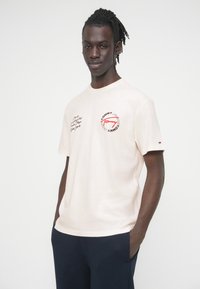 Μπεζ βαμβακερό T-shirt με στρογγυλή λαιμόκοψη, κοντά μανίκια και κεντητά κόκκινα και μαύρα λογότυπα στο στήθος και το μανίκι.