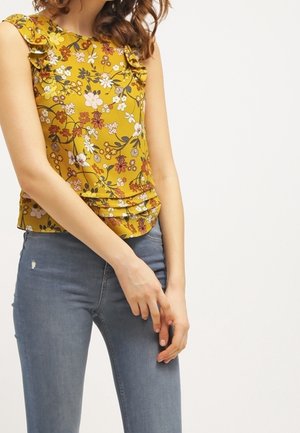 Mujer con una blusa sin mangas de flores color amarillo mostaza con volantes en los hombros y jeans ajustados azules, frente a un fondo liso.