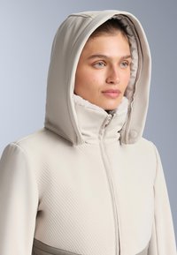 Hellbeige Jacke mit gepolsteter Kapuze, die einen strukturierten oberen Bereich und einen glatten unteren Abschnitt aufweist. Reißverschluss vorne und weiche Futterakzente.