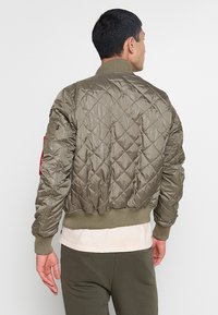 Olivgrüne, gesteppte Bomberjacke mit gerippten Bündchen und Saum, die einen roten Akzent am linken Arm aufweist und aus leichtem, glänzendem Material gefertigt ist.