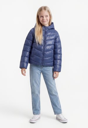 Niña joven con cabello rubio largo, que lleva una chaqueta acolchada azul, vaqueros azul claro y zapatillas blancas, de pie y sonriendo.