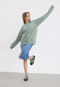 Sweatshirt surdimensionné vert menthe, jupe midi en denim avec fente sur le côté, baskets bleu marine avec accents verts, mannequin dans une pose active, arrière-plan minimal.