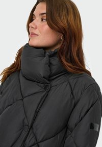 Juodas pūkinis švarkas su aukštu apykakle, turintis deimanto formos siuvinėjimą, didelius mygtukus ir lygią medžiagos tekstūrą.