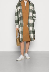 Grön och vit rutig oversized kappa över en brun klänning, i kombination med vita sneakers och en ljusbrun väska. Släta texturer och en avslappnad passform.