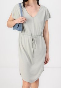 Helles graues Kleid mit V-Ausschnitt, kurzen Ärmeln und elastischem Bund. Es verfügt über einen Kordelzug und seitliche Schlitze für bessere Bewegungsfreiheit.