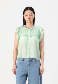 Blusa sem mangas de tom verde claro, com detalhes em folhos, frente com botões e tecido texturizado. Combinada com jeans de corte largo.