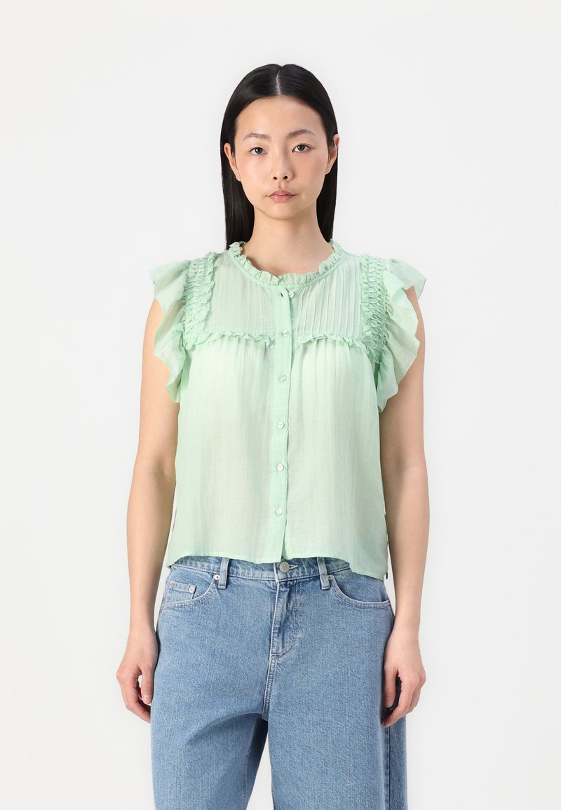Blusa sem mangas de tom verde claro, com detalhes em folhos, frente com botões e tecido texturizado. Combinada com jeans de corte largo.