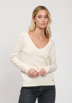 Lyshåret kvinde iført en cremefarvet V-hals sweater og mørke jeans, stående mod en ensartet lysegrå baggrund.