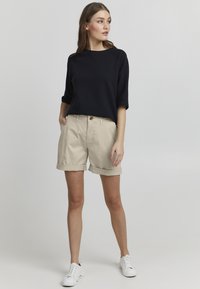 Pull en coton noir à manches courtes, associé à un short en coton beige avec des ourlets roulés et une fermeture à bouton. Des baskets blanches complètent le look.