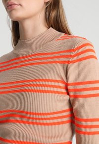 Sweter z prążkowanej dzianiny w kolorze beżowym z odważnymi pomarańczowymi paskami. Posiada wysoki, dopasowany dekolt oraz teksturowaną tkaninę z przylegającym krojem.