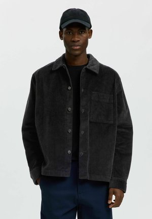 Homme portant une veste noire en velours côtelé boutonnée, une chemise noire, un pantalon bleu marine et une casquette de baseball noire, se tenant devant un fond clair uni.