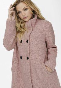 Manteau texturé rose avec un col montant, des boutons noirs à double boutonnage et des poches latérales. Matière douce avec une coupe décontractée.