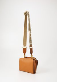 Brun skinn crossover-veske med teksturert overflate, med gyllen glidelås og en beige stropp med logo-accents. Kompakt rektangulær form.