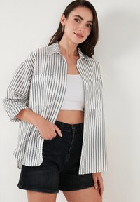 Striped button-up shirt in white and navy, featuring long sleeves, chest pockets, and a relaxed fit, paired with black denim shorts.