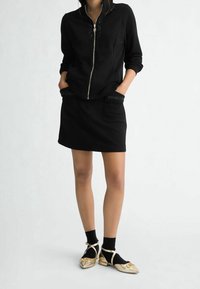 Giacca nera con cerniera e dettagli borchiati, abbinata a una minigonna nera. Ballerine dorate indossate con calzini neri per un contrasto evidente.