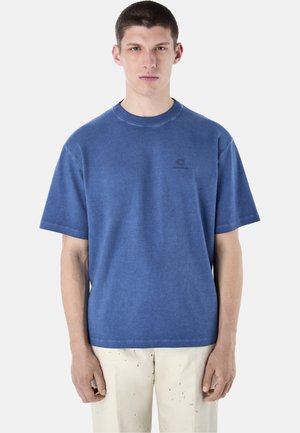 Młody mężczyzna ubrany w niebieską koszulkę z krótkim rękawem i jasnobeżowe spodnie z drobnymi ciemnymi wzorami, stojący na jednolitym tle.