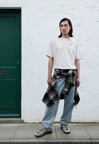 Smėlio spalvos marškinėliai, šviesiai mėlyni platūs džinsai, juoda ir pilka languota marškinėliai pririšti aplink liemenį, ir marginti sportbačiai. Fone žalia durys.