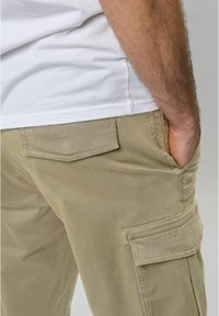 Khaki spodnie cargo wykonane z mieszanki bawełny, z bocznymi kieszeniami i luźnym krojem. Projekt zawiera widoczne szwy i gładką teksturę.