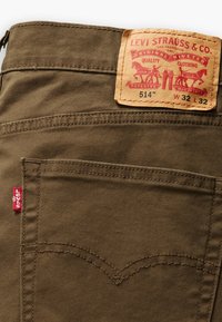 Brown cotton trousers with a leather brand patch, red tab, and curved stitching details on the back pockets. Size label: W32 L32.