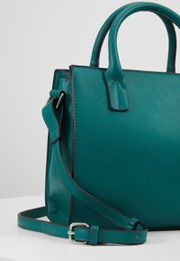 Sac à main en cuir turquoise avec doubles poignées supérieures et bandoulière ajustable, posé sur une surface blanche sur un fond clair.