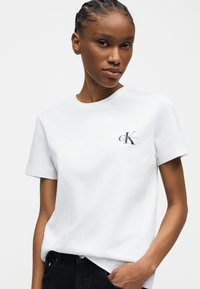 Γυναίκα με πλεξούδες, ντυμένη με λευκό κοντομάνικο T-shirt Calvin Klein και μαύρο παντελόνι, ποζάρει με το ένα χέρι στην τσέπη.