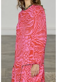 Robe rose vif avec des motifs abstraits rouges audacieux, manches longues et poignets froncés. Caractérisée par un col rond et une silhouette fluide.