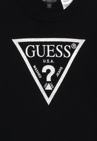 Černá mikina s velkým kovovým stříbrným trojúhelníkovým logem, na jehož vrcholu je nápis "GUESS", ve středu otazník a text pod ním.