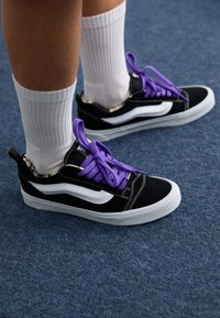Sneakers in suede nero con dettagli bianchi e lacci viola. Presentano una tomaia texture e una suola in gomma bianca. Indossati con calze bianche a coste.