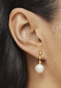 Pendientes de oro con tres elementos esféricos: dos pequeñas bolas de oro arriba y una perla blanca más grande en la parte inferior, llevados en una oreja perforada.