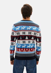 Homme portant un pull de Noël bleu, rouge, noir et blanc avec des motifs de rennes, de flocons de neige et de sapins, assorti à un jean bleu, de dos.